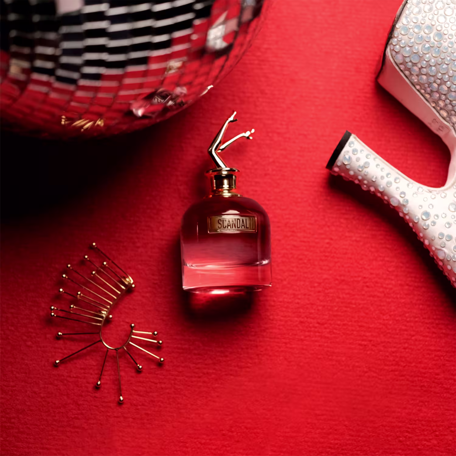 Perfume em frasco de vidro vermelho com a palavra "Scandal" gravada.  Ao redor, há acessórios decorativos metálicos dourados em formato de espiral e estrela. Há também uma embalagem vermelha com detalhes perolados.