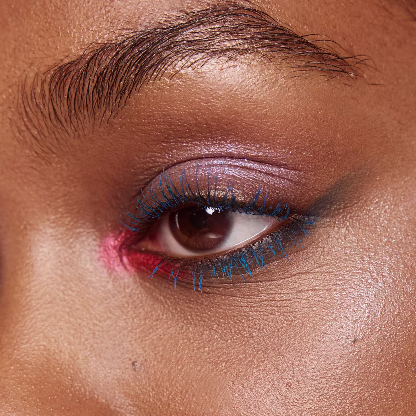 A foto mostra um rosto feminino aproximado, com foco no olho castanho. A pálpebra está pintada com uma sombra  rosa claro e os cílios bem definidos e alongados na cor azul.