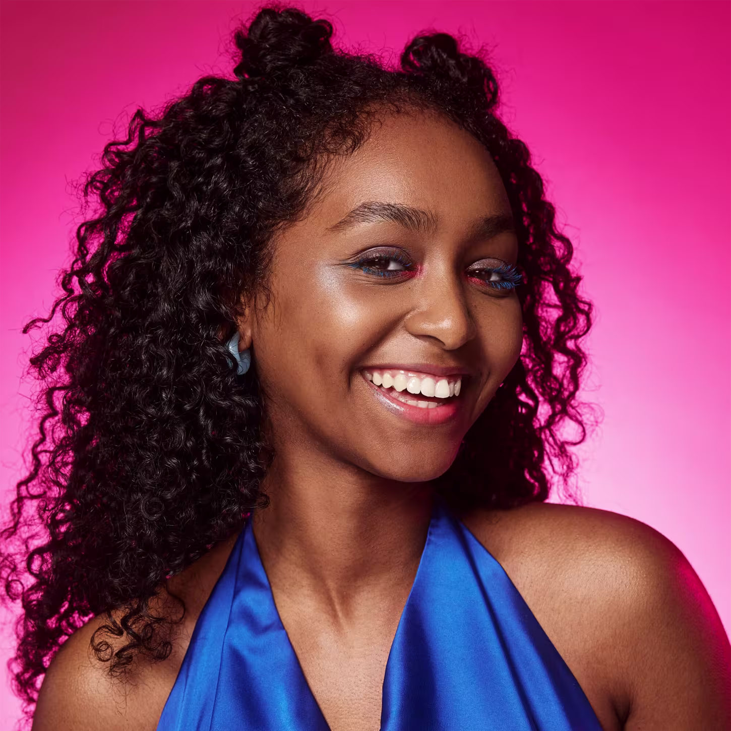 Esta imagem apresenta uma mulher negra sorridente com cabelos cacheados e volumosos em um fundo rosa vibrante. Ela está usando um top azul Seu sorriso largo exibe dentes brancos brilhantes.