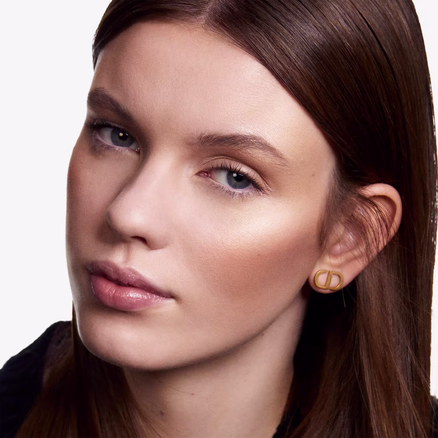 A imagem mostra uma mulher jovem com cabelos castanhos escuros e pele clara. Ela está usando maquiagem natural, com sombra marrom suave nos olhos, delineador preto discreto e batom rosa claro brilhante nos lábios cheios.