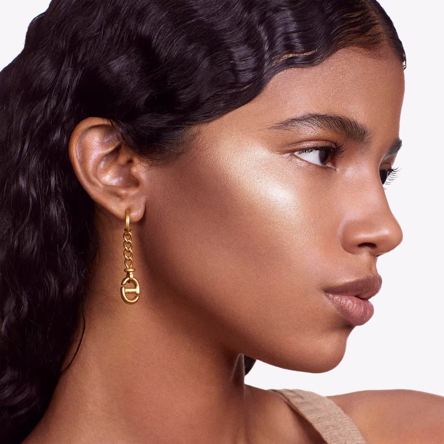 Esta é uma imagem de uma mulher com pele bronzeada e traços delicados, promovendo um produto de beleza. Ela tem cabelos escuros e ondulados, olhos bem delineados com maquiagem e lábios levemente rosados. Usa um brinco dourado com um design de elos.