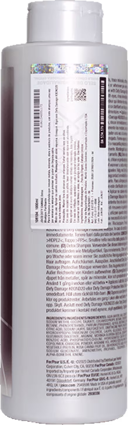Frasco cilíndrico branco de plástico para shampoo da marca Joico. O rótulo contém informações detalhadas sobre o produto, incluindo ingredientes e instruções, em várias línguas. O frasco tem tampa prata e apresenta as cores branca e roxa.