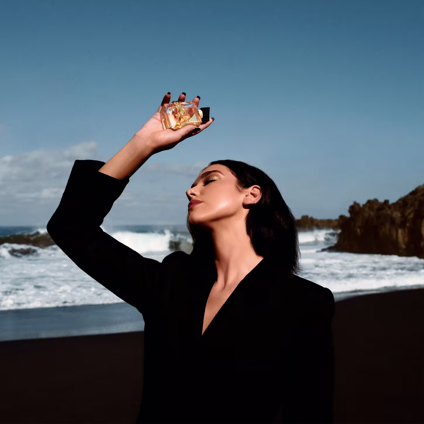 A imagem mostra a cantora Dua Lipa, uma mulher jovem branca de cabelos pretos curtos, segurando o perfume. Ela está com um terno preto e ao fundo é possível ver uma praia.