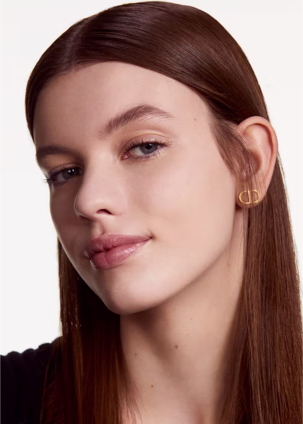 Esta imagem apresenta uma mulher jovem com longos cabelos castanhos lisos e olhos claros. Ela usa brincos dourados e tem uma expressão serena, com lábios rosados e maquiagem suave.