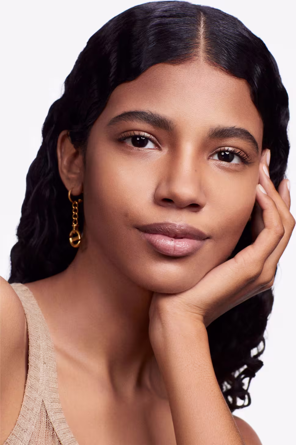Esta imagem apresenta uma modelo jovem com traços étnicos. Ela tem pele bronzeada, olhos castanhos marcantes e lábios cheios e rosados. Seus cabelos são negros e ondulados. Ela usa brincos dourados e toca suavemente o rosto com uma das mãos.