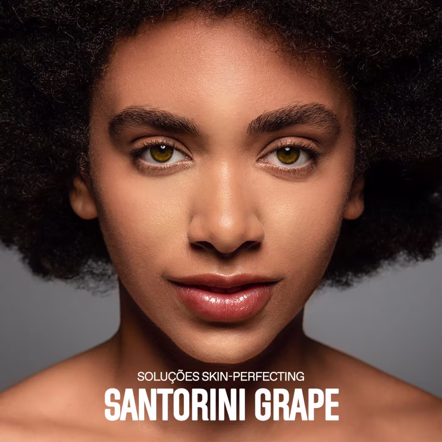 A imagem mostra um close-up do rosto de uma mulher negra com cabelos crespos e curtos. Ela tem olhos amendoados de cor amarelo-esverdeada. Sua pele tem um aspecto aveludado e uniforme, provavelmente resultado do uso dos produtos anunciados.