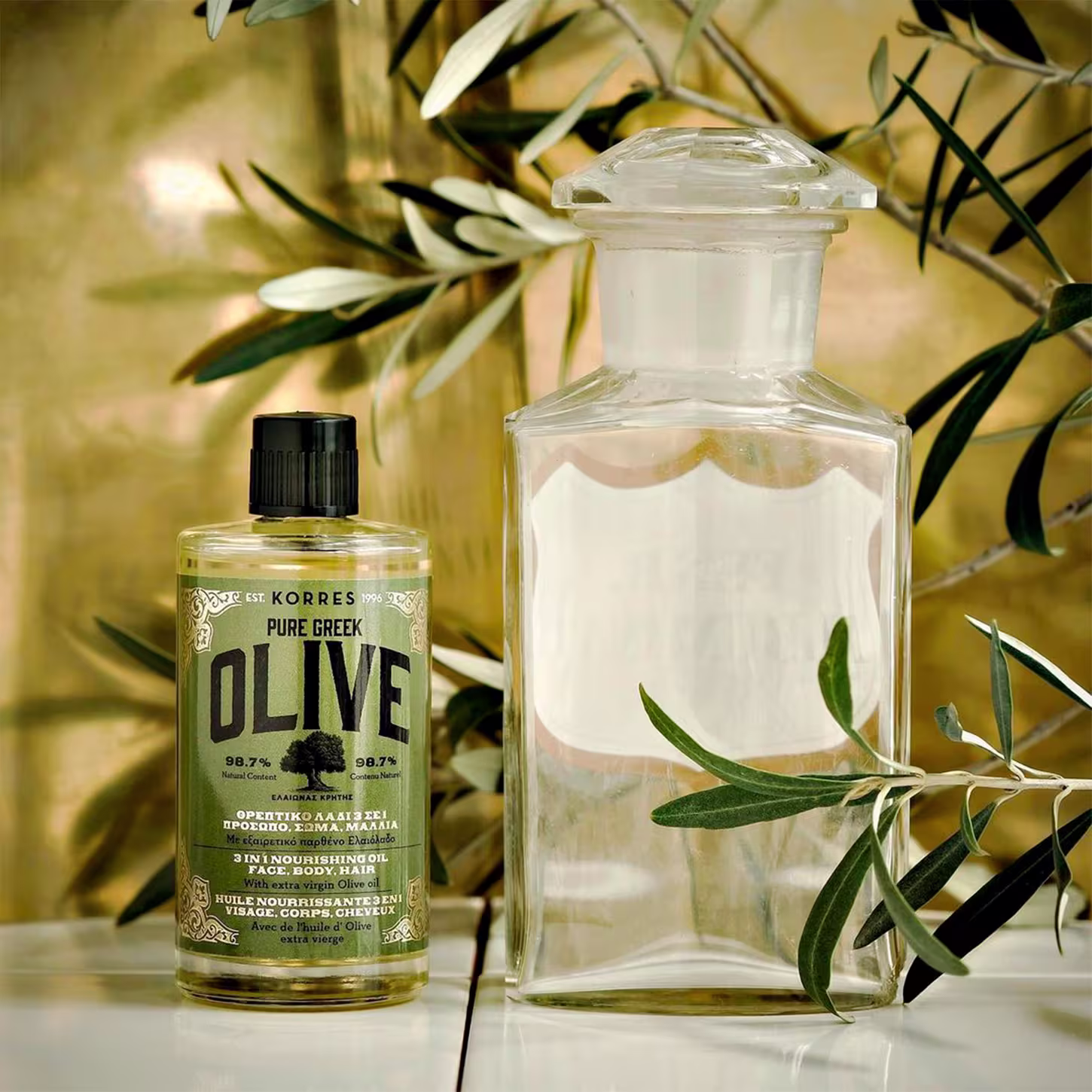 A imagem mostra o produto e ao lado um frasco de vidro transparente. Ambos apresentam galhos de oliveira ao redor, simbolizando os ingredientes naturais. As cores predominantes são transparente, verde oliva e preto.