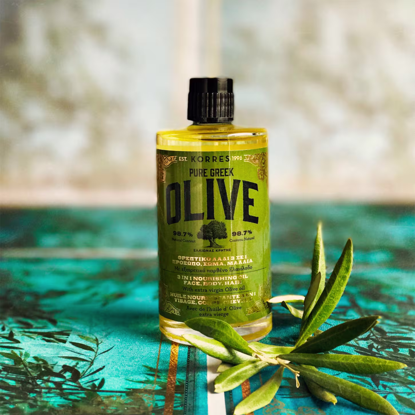 A imagem mostra o frasco acompanhado de um ramo de oliveira com folhas verdes e amarelas sobre uma superfície azul turquesa, criando um contraste vibrante.