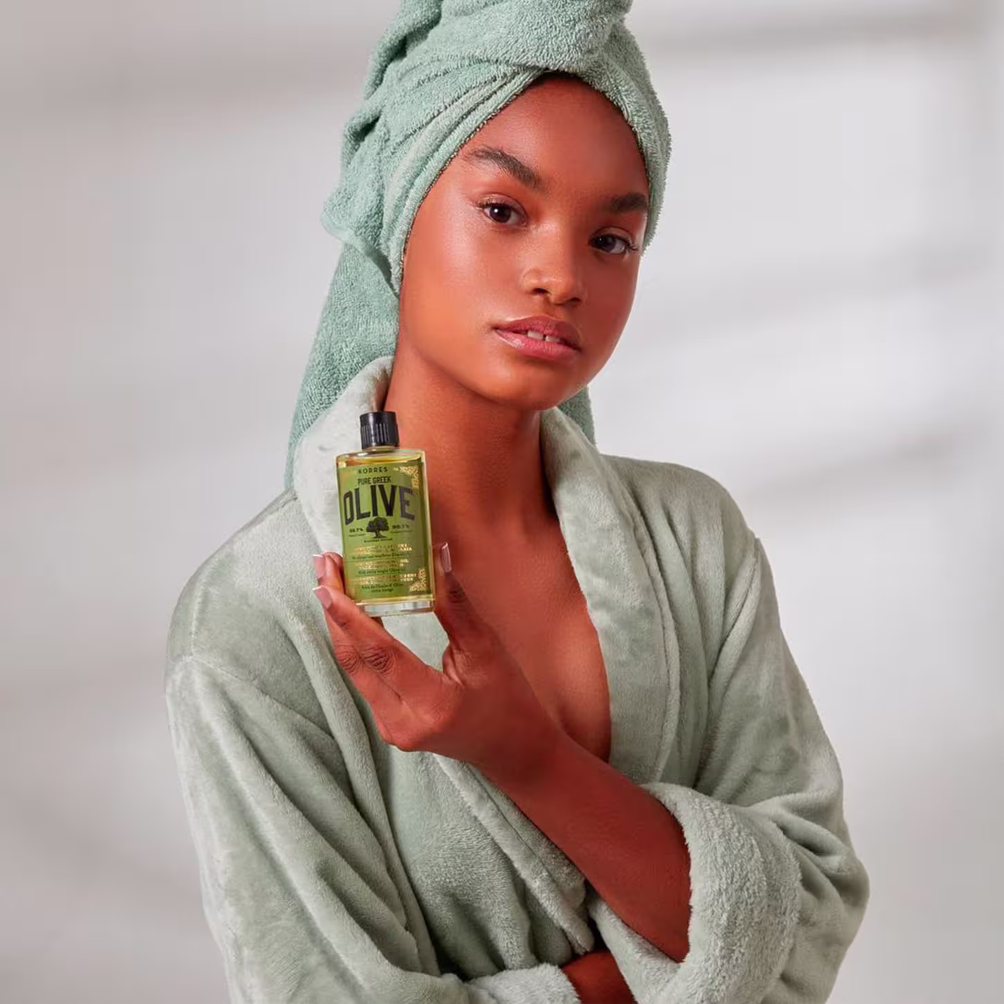 A mulher mostra uma mulher de pele negra com roupão e toalha na cabeça. Ela está com uma expressão serena e segura o frasco em uma das mãos.