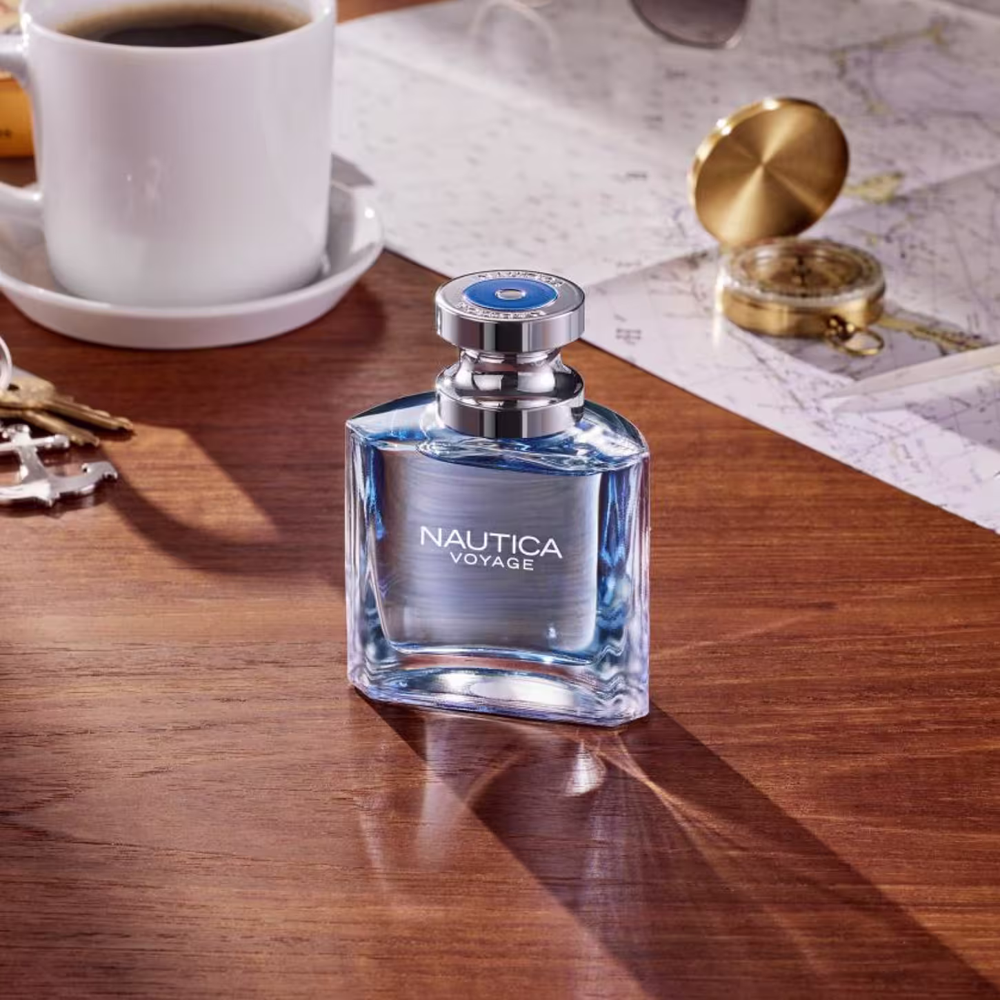Perfume Nautica Voyage em frasco retangular de vidro azul transparente com tampa cromada. Está sobre uma superfície de madeira, acompanhado de uma xícara de café branco e uma bússola dourada.