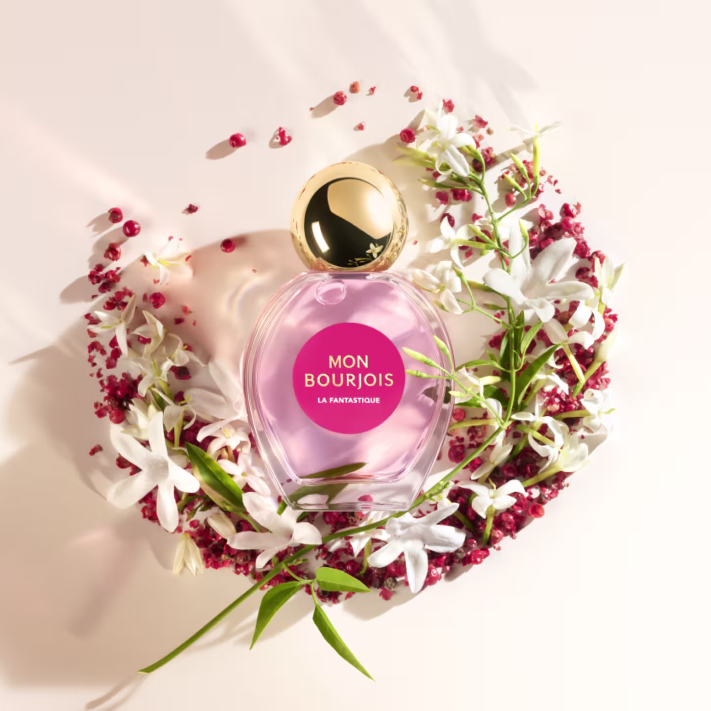 cercado por flores brancas e vermelhas, com pequenos detalhes vermelhos espalhados ao redor. Embalagem apresenta cores rosa, branca, verde e dourada, com destaque para o frasco de perfume centralizado.