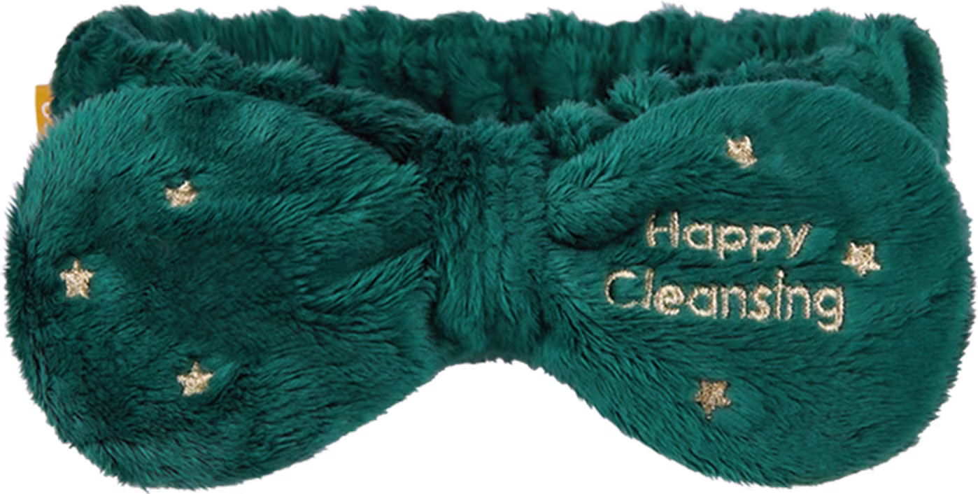O produto apresentado é uma faixa de cabelo felpuda da marca Banila Co. Possui cor verde-esmeralda, com pequenos detalhes dourados em forma de estrelas. A embalagem exibe o texto "Happy Cleansing" bordado em dourado.
