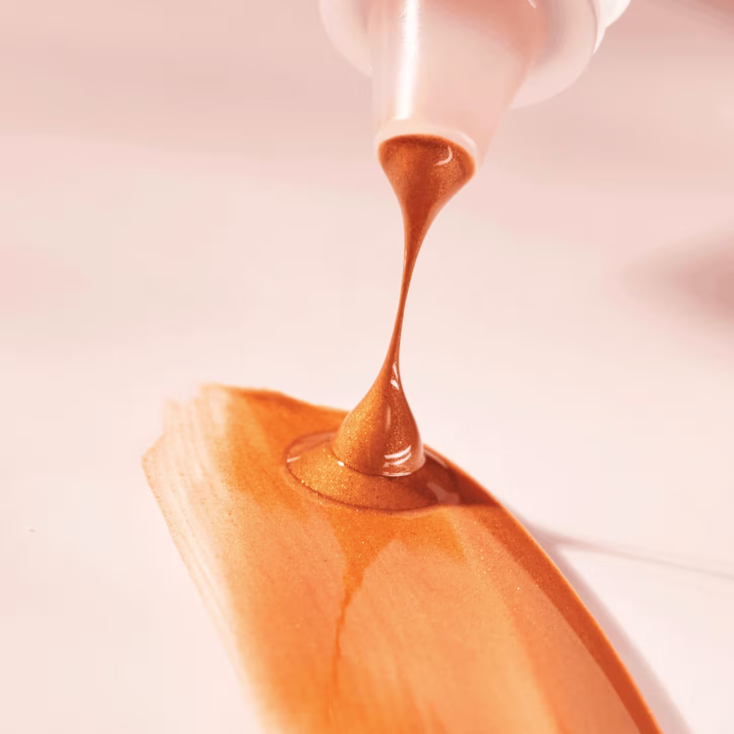 O bronzer está sendo despejado de um frasco ou recipiente, formando um filete ou gota alongada e pontiaguda. Destacando sua textura sedosa e brilhante contra um fundo desfocado em tons claros e suaves.