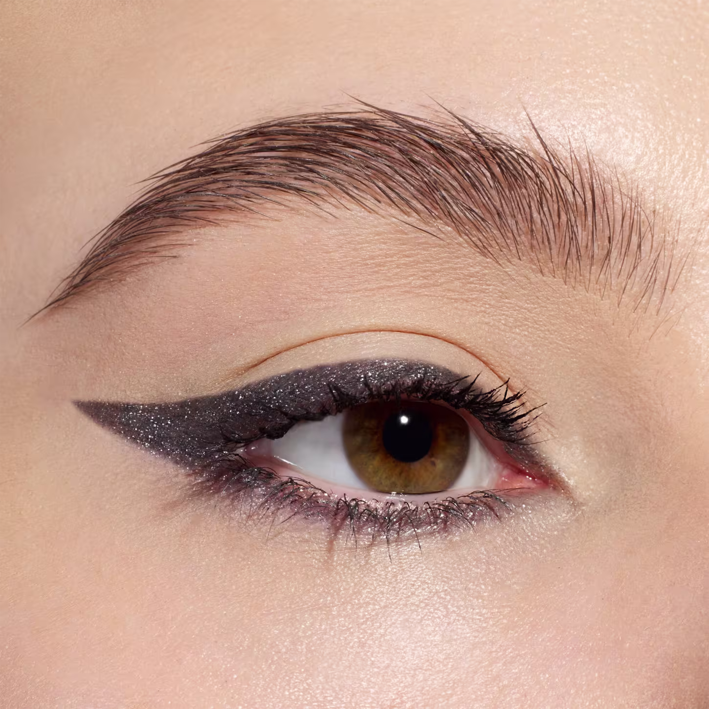 A imagem mostra uma maquiagem de olhos com um delineado gatinho dramático em glitter chumbo cintilante. O delineado é alongado e afiado nas extremidades externas, criando um efeito marcante. A sombra é suave e brilhante, realçando os olhos castanhos.