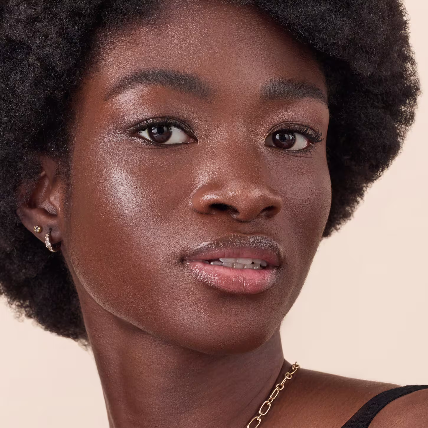 Jovem mulher negra com cabelos crespos e curtos. Ela tem olhos castanhos expressivos, sobrancelhas bem delineadas e lábios cheios levemente brilhantes. Sua pele é aveludada e brilhante, com um tom marrom profundo e saudável.