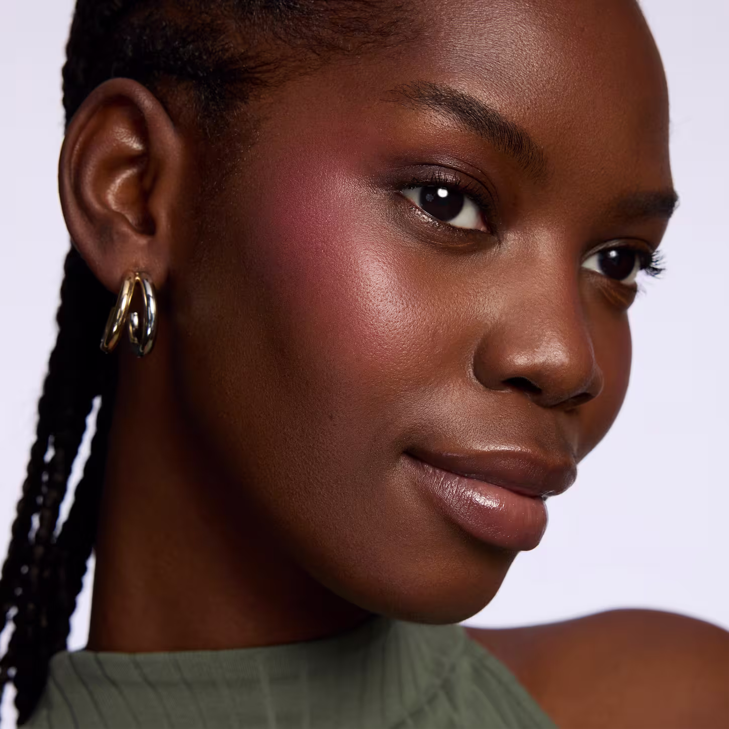 Esta imagem mostra um close-up do rosto de uma mulher negra jovem com traços delicados e pele brilhante. Ela tem olhos castanhos profundos, sobrancelhas bem delineadas e lábios cheios e brilhantes. Seus cabelos negros estão trançados em finas tranças.