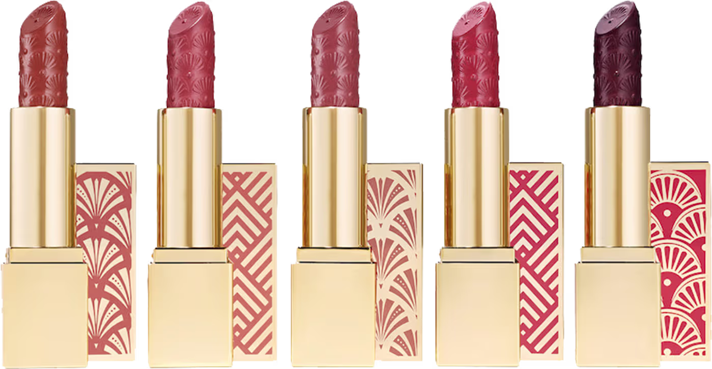 Cinco batons Estée Lauder. Os batons têm um formato cilíndrico tradicional, com uma capa dourada metálica na parte inferior. As cores variam de rosa claro a rosa escuro.