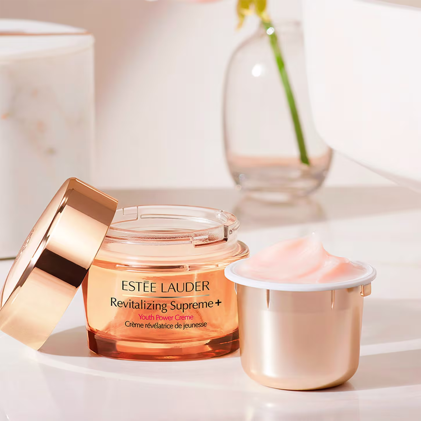 Produto de cuidados com a pele Estée Lauder. Frasco contendo um creme facial amarelo-alaranjado chamado "Revitalizing Supreme+ Youth Power Creme". Uma flor amarela em um vaso de vidro transparente complementa a composição.