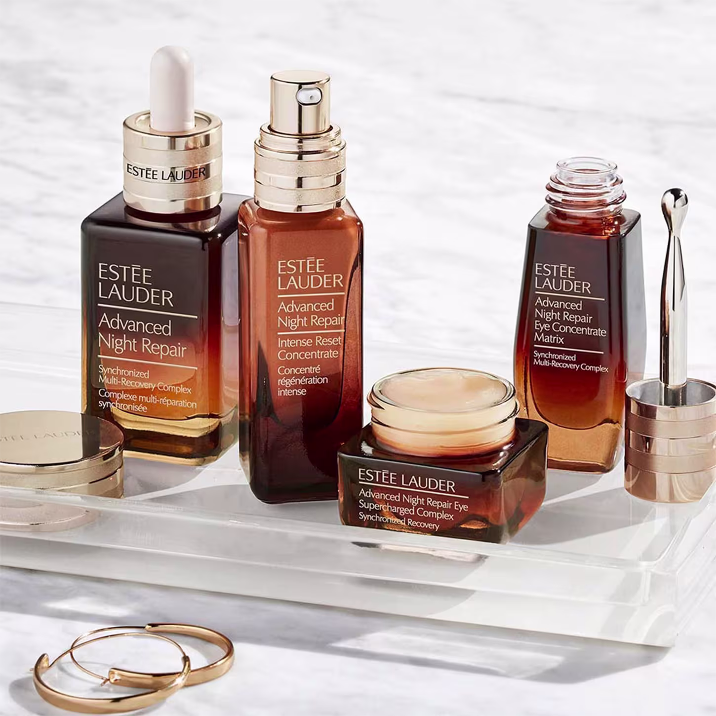 A imagem apresenta uma variedade de produtos de cuidados com a pele Estée Lauder. Há frascos séruns ou concentrados, com rótulos que indicam serem da linha "Advanced Night Repair". Também há um frasco menor de creme para a área dos olhos da mesma linha.