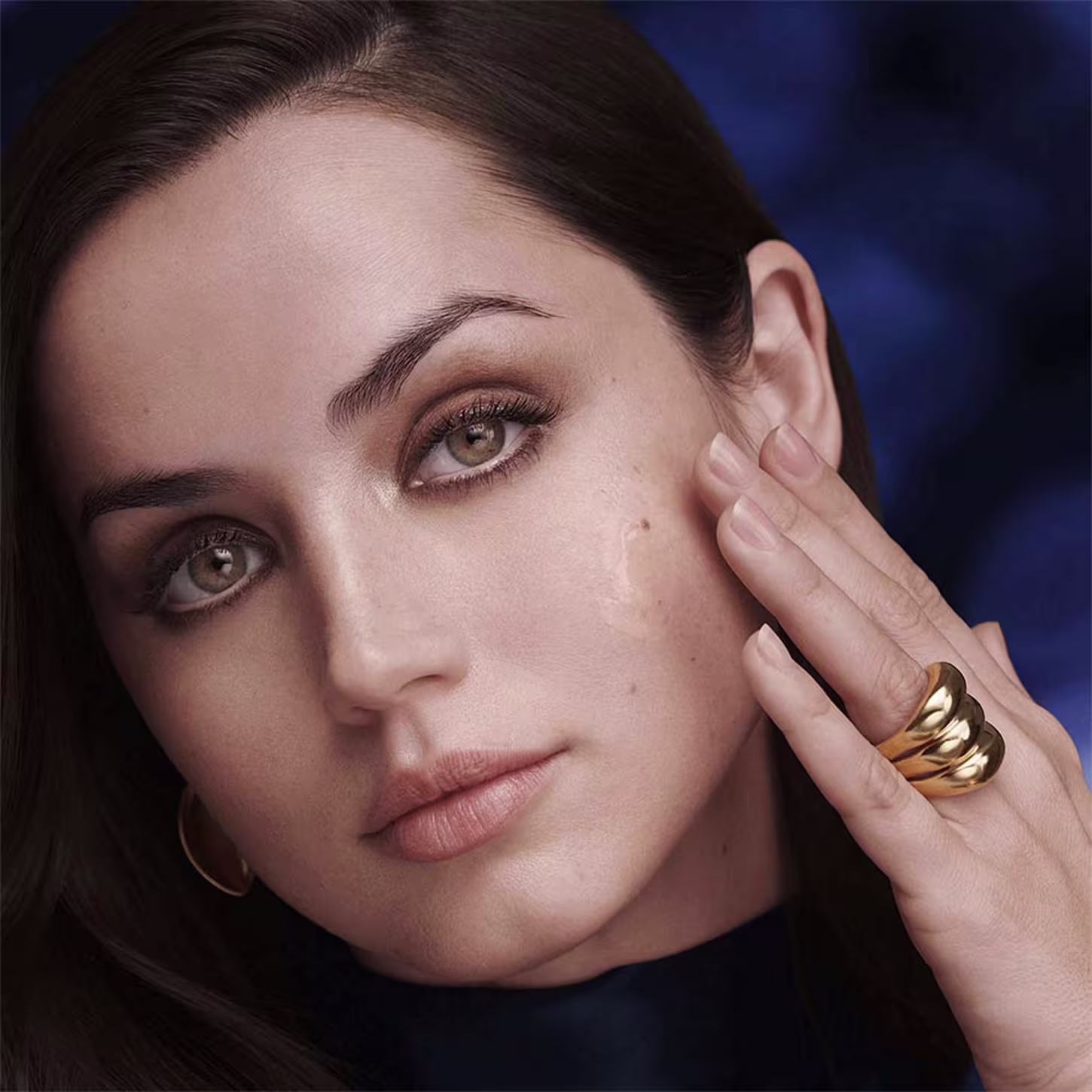 Modelo feminina com aparência sofisticada e maquiagem impecável. A modelo toca seu rosto delicadamente com uma das mãos, onde usa um anel dourado enrolado. O fundo é azul escuro, criando um contraste elegante com sua pele clara e traços delicados.