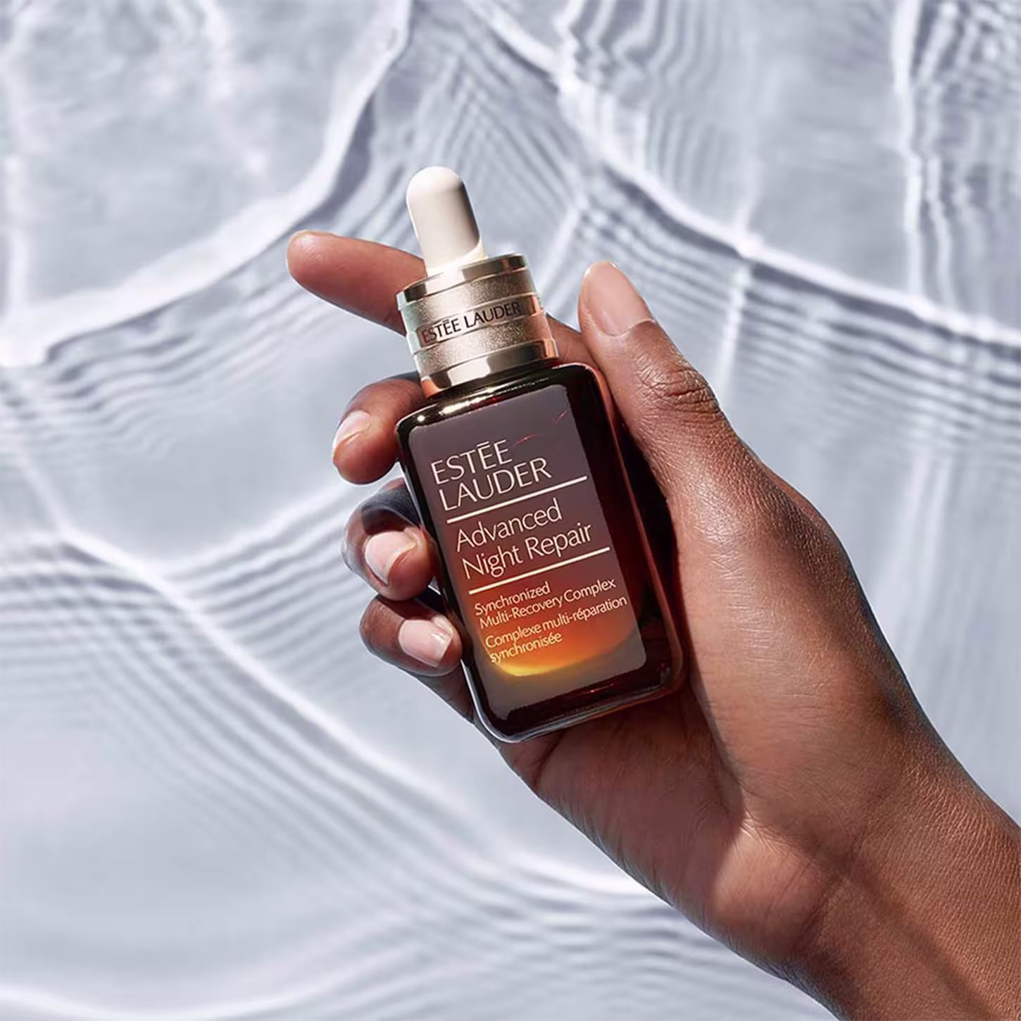 Sérum facial Estée Lauder. Rótulo com o nome "Advanced Night Repair" e descrição "Synchronized Multi-Recovery Complex". Embalagem segurada por uma mão de pele escura, com unhas pintadas, em um fundo desfocado com padrões ondulados em tons claros.
