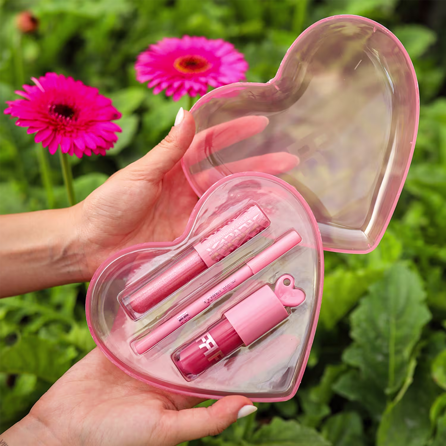 Conjunto de produtos de beleza da marca Fran by Franciny Ehlke em embalagem em formato de coração, com tons de rosa, em um ambiente externo com flores gerbera rosa ao fundo.