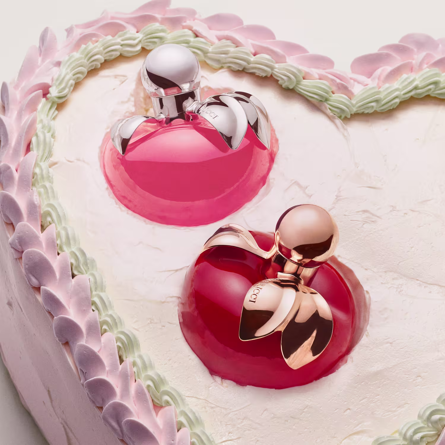 Dois frascos de perfume feminino com embalagens em tons de rosa e vermelho, com detalhes em metal prateado. O frasco maior possui um laço decorativo na tampa. Ambos os produtos exibem um design elegante e sofisticado e estão no topo de um bolo.