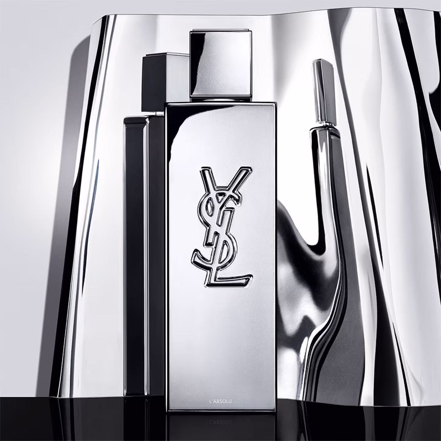 Frasco de perfume de design minimalista prateado, com o logotipo da marca Yves Saint Laurent em destaque. A embalagem possui uma forma geométrica simples e elegante, com uma tampa destacada no topo.