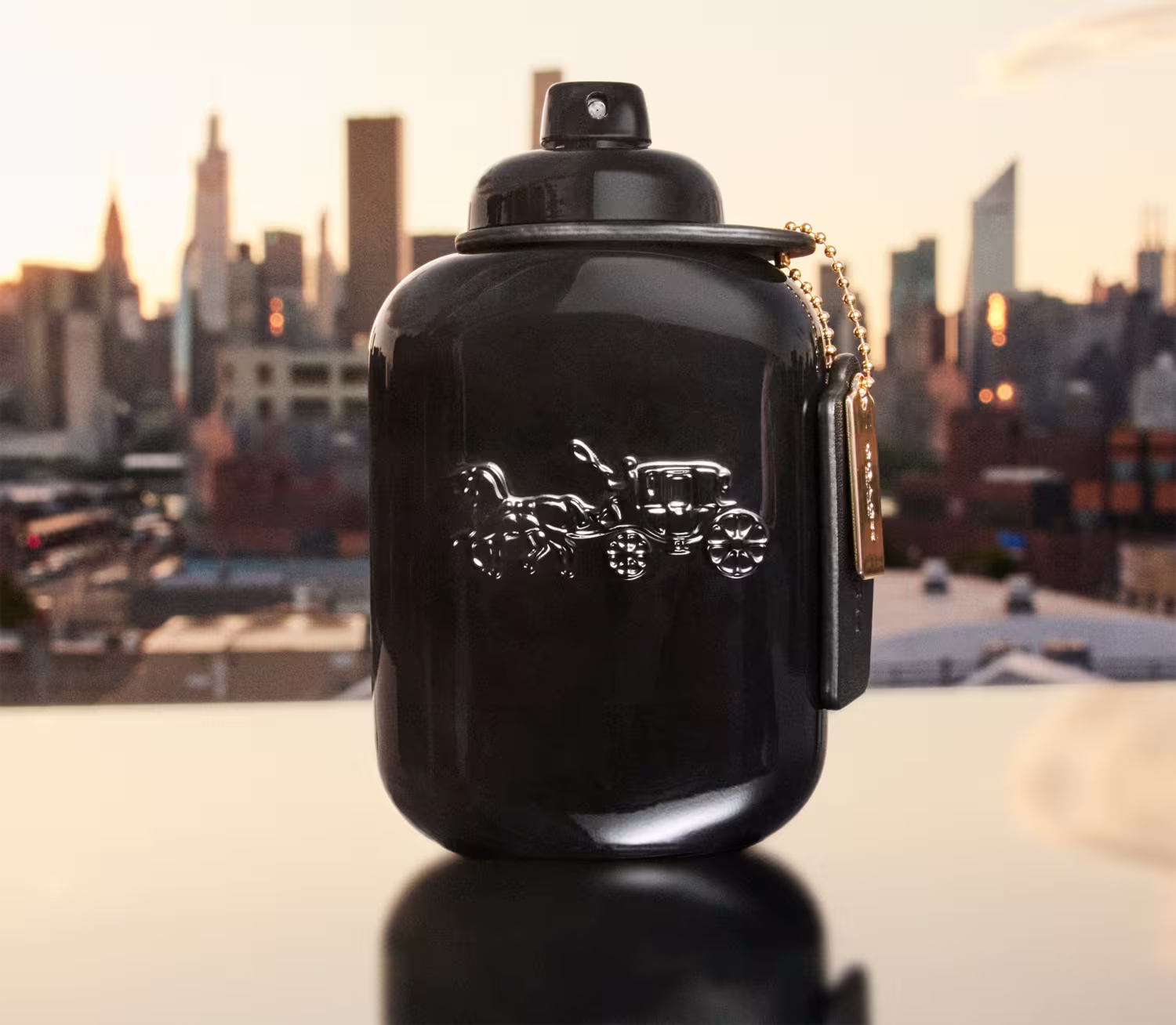 O produto de beleza da marca Coach é apresentado em um cenário urbano com arranha-céus ao fundo, sugerindo um ambiente cosmopolita e moderno. A embalagem preta e o design elegante do frasco destacam-se contra o pano de fundo da cidade.