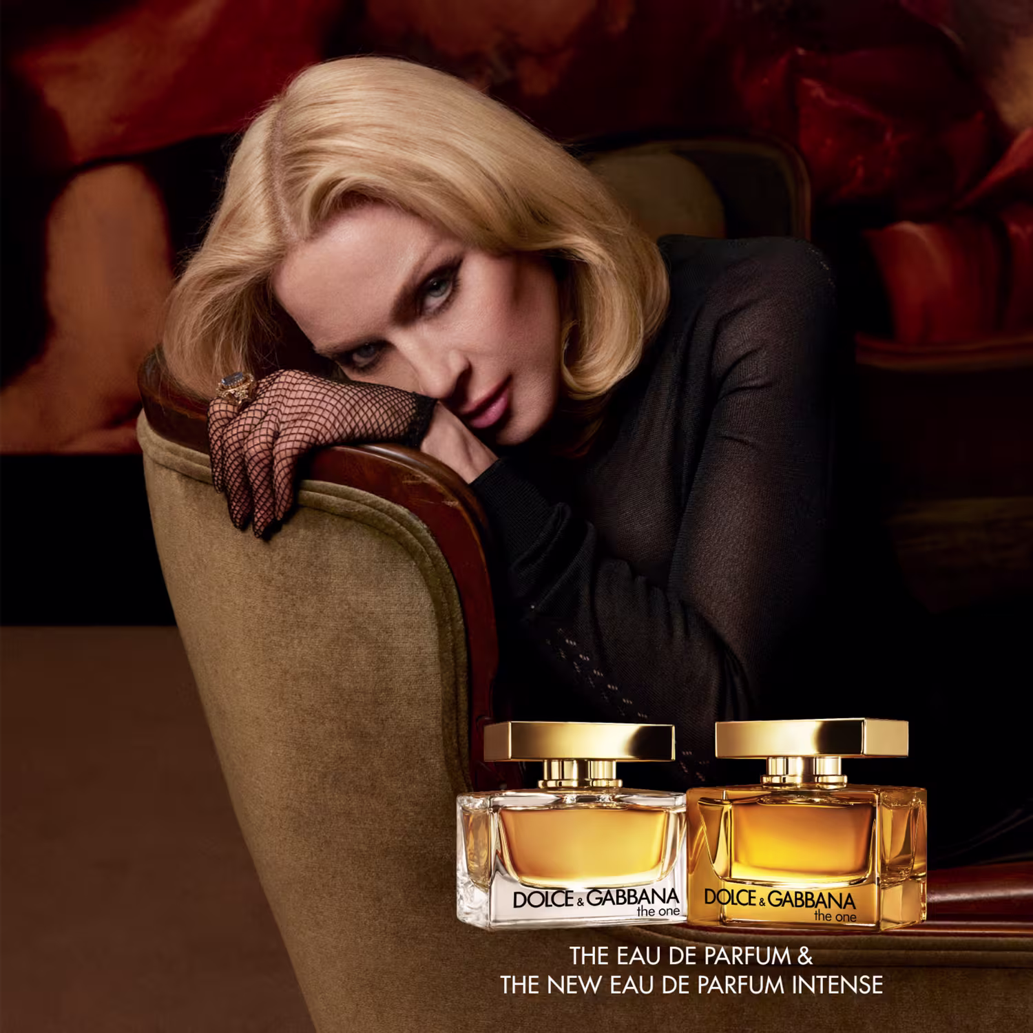 Uma mulher loira vestida de preto está reclinada em uma cadeira, com os braços apoiados. Ela usa luvas de renda preta e está rodeada por três frascos de perfume da marca Dolce & Gabbana, em um ambiente escuro e dramático.