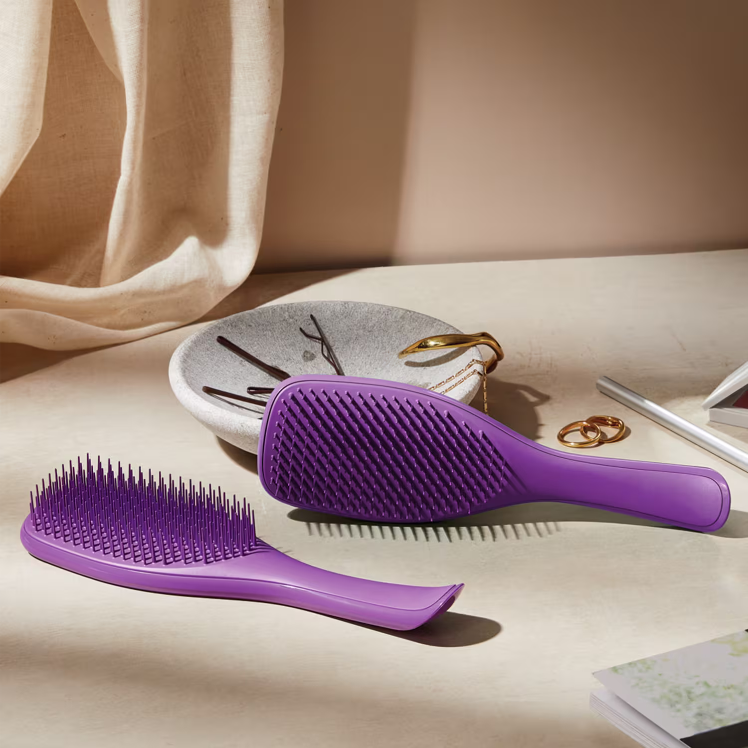 Ambiente de penteado e cuidados pessoais com acessórios em tons neutros, destacando-se a escova de cabelo da marca Tangle Teezer em cor roxa, posicionada em primeiro plano.