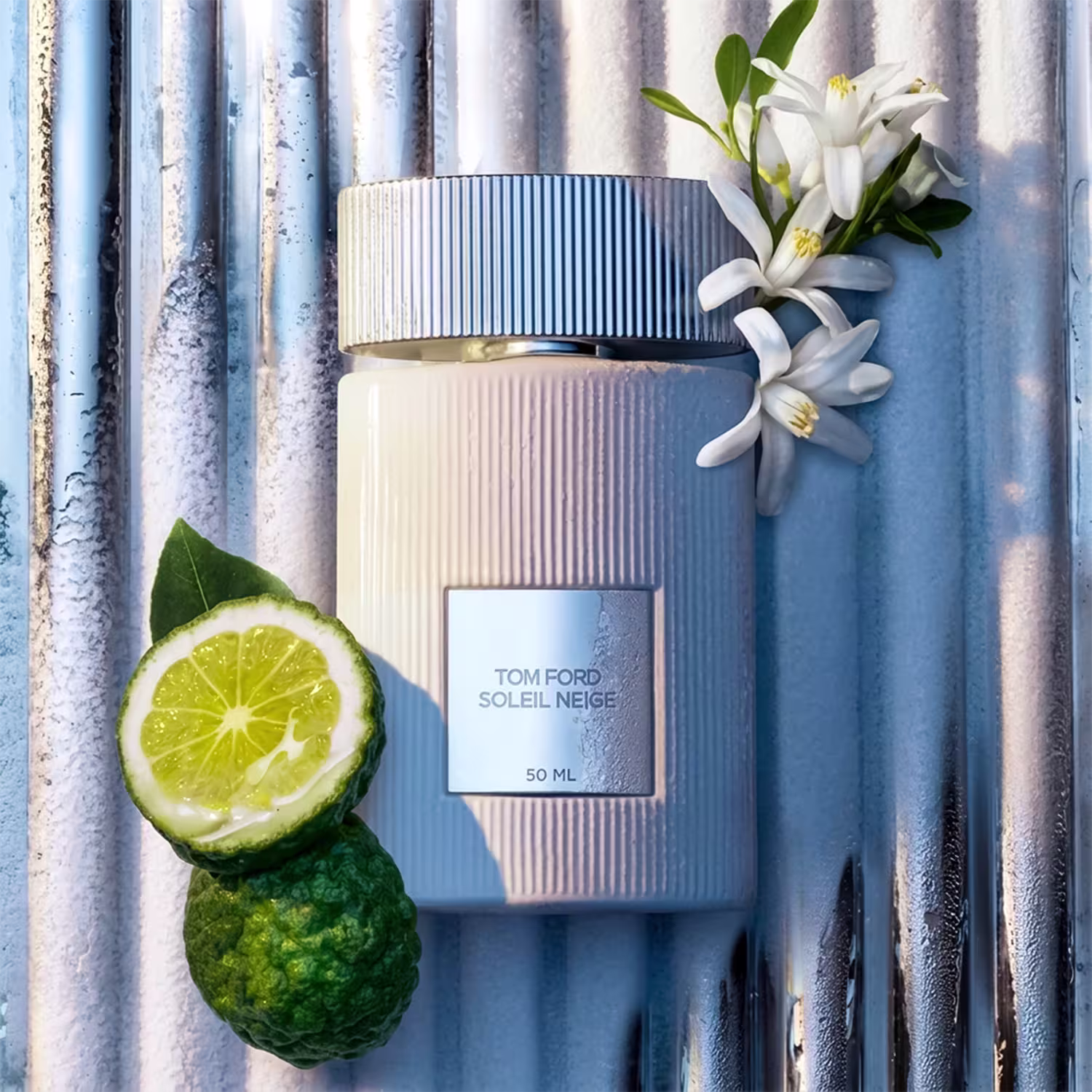 Frasco de perfume Tom Ford Soleil Neige, com embalagem branca e detalhes em azul, acompanhado de flores brancas e uma fatia de limão verde.