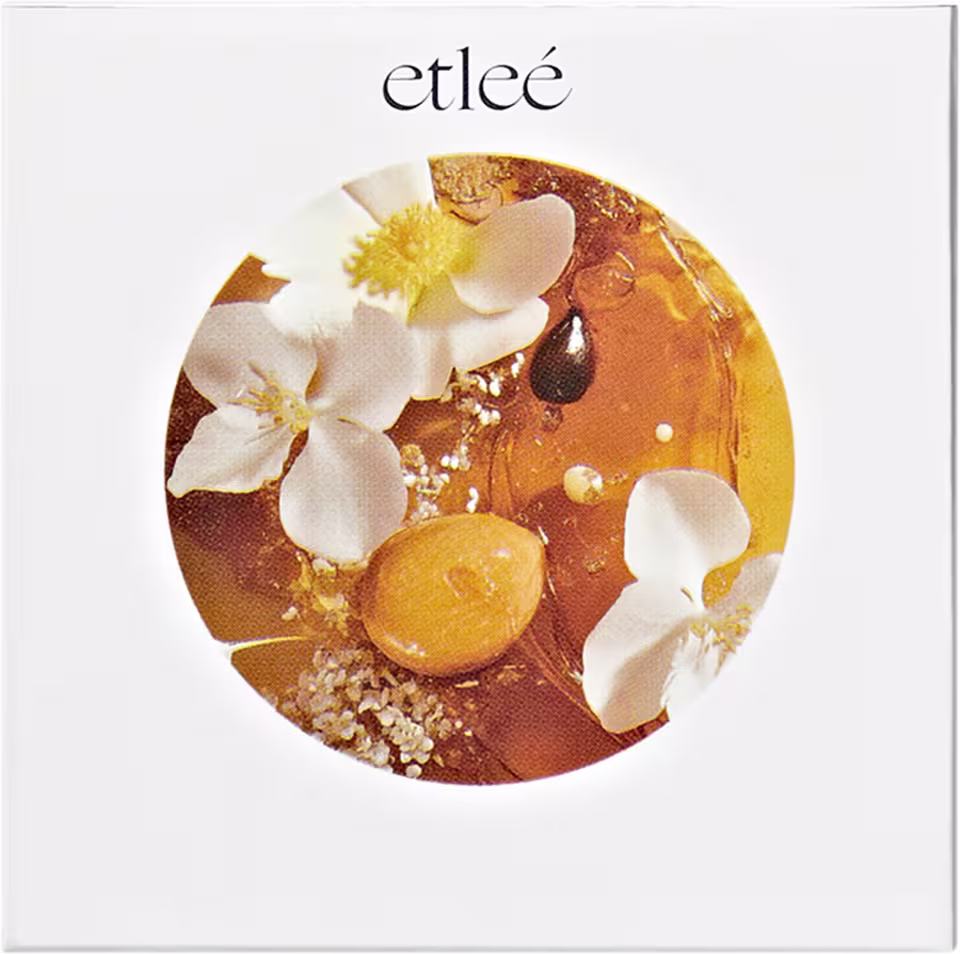 Caixa branca com logotipo Etleé e imagem circular  com flores brancas, gotas douradas de caramelo e pistache sugerindo a composição olfativa do perfume.