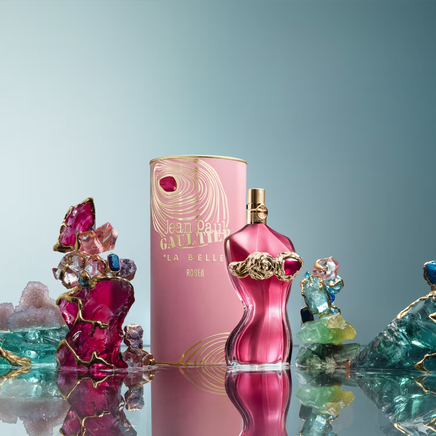 Ambiente com perfume da marca Jean Paul Gaultier, rodeado por cristais, pedras preciosas e flores em tons vibrantes de rosa, roxo e vermelho