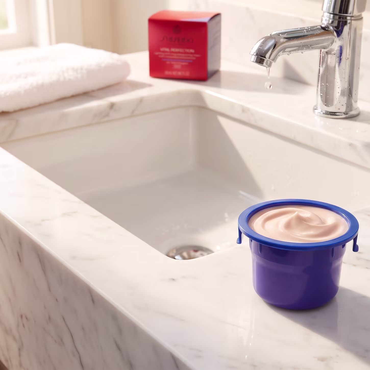 Ambiente de banheiro com pia de mármore, torneira cromada e caixa vermelha da marca Shiseido. Ao lado, um recipiente azul contendo refil do Shiseido Vital Perfection Uplifting and Firming Day Cream SPF 30