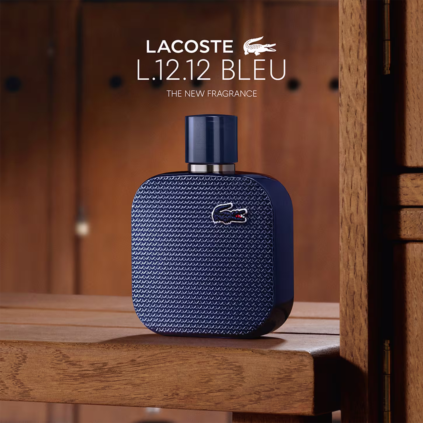 Frasco de perfume Lacoste de cor azul escuro, com textura de grade e logotipo da marca em destaque. Embalagem cilíndrica com tampa preta, apresentando design elegante e sofisticado.