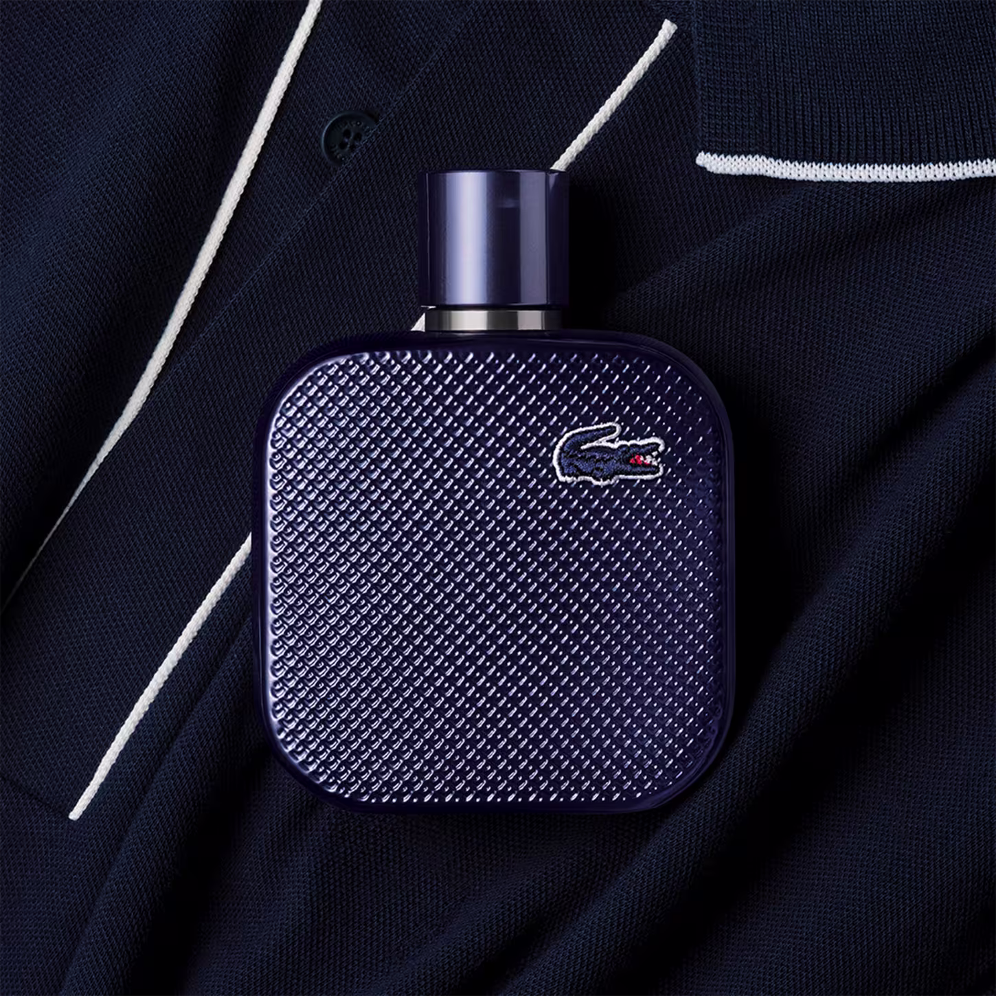 Frasco de perfume Lacoste de cor azul escuro, com textura de grade e logotipo da marca em destaque. Embalagem cilíndrica com tampa preta, apresentando design elegante e sofisticado.