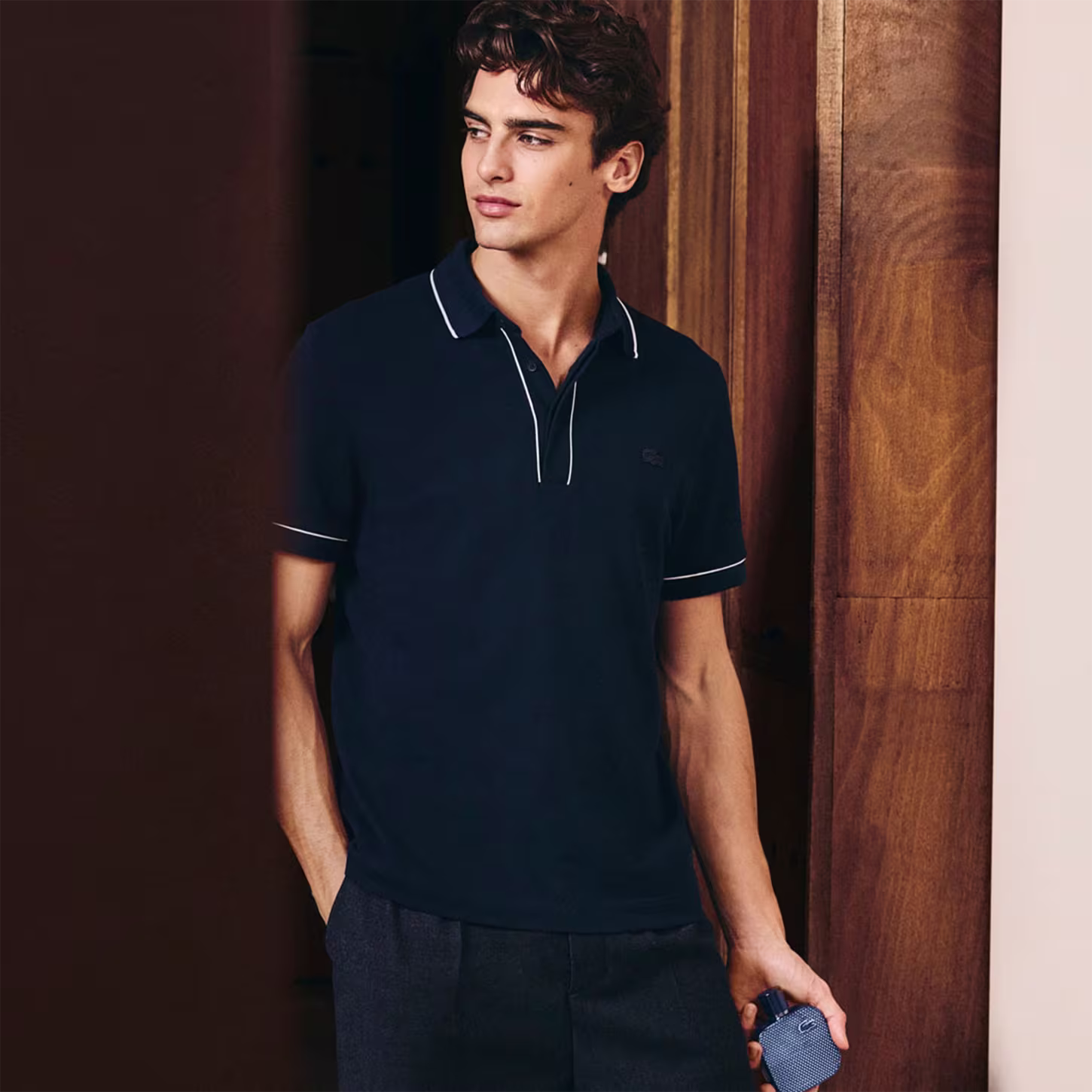 Frasco de perfume Lacoste de cor azul escuro, com textura de grade e logotipo da marca em destaque. Embalagem cilíndrica com tampa preta, apresentando design elegante e sofisticado.