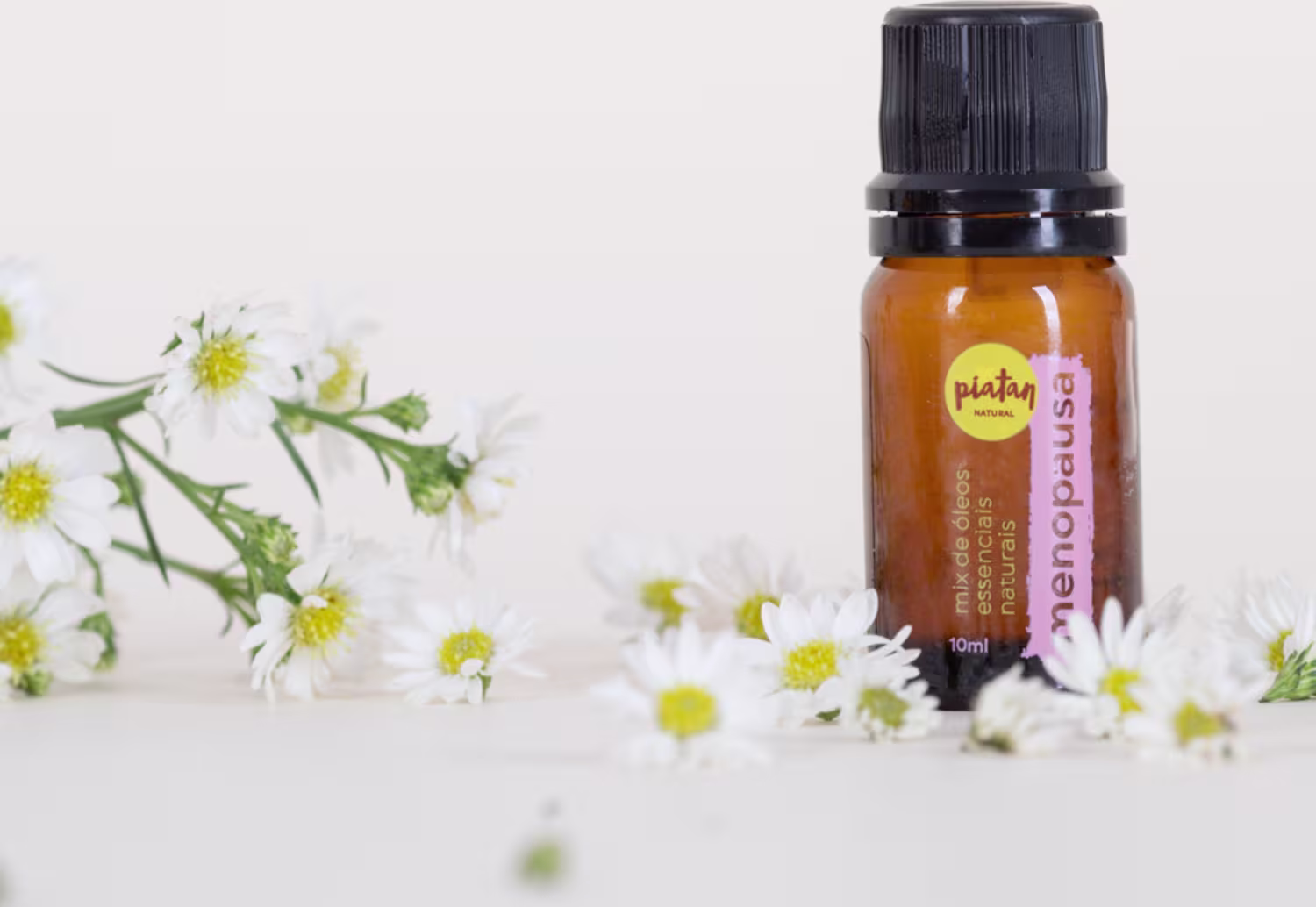 O frasco está posicionado ao lado de pequenas flores brancas com centros amarelos, provavelmente camomilas, espalhadas ao redor, criando um cenário natural e orgânico.