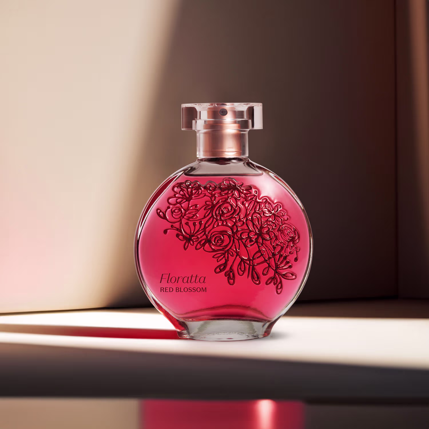 O frasco é transparente com um design elegante e arredondado. O líquido perfumado possui uma cor rosa vibrante e no frasco é visível uma decoração delicada com desenhos de rosas e galhos em um tom vermelho escuro.