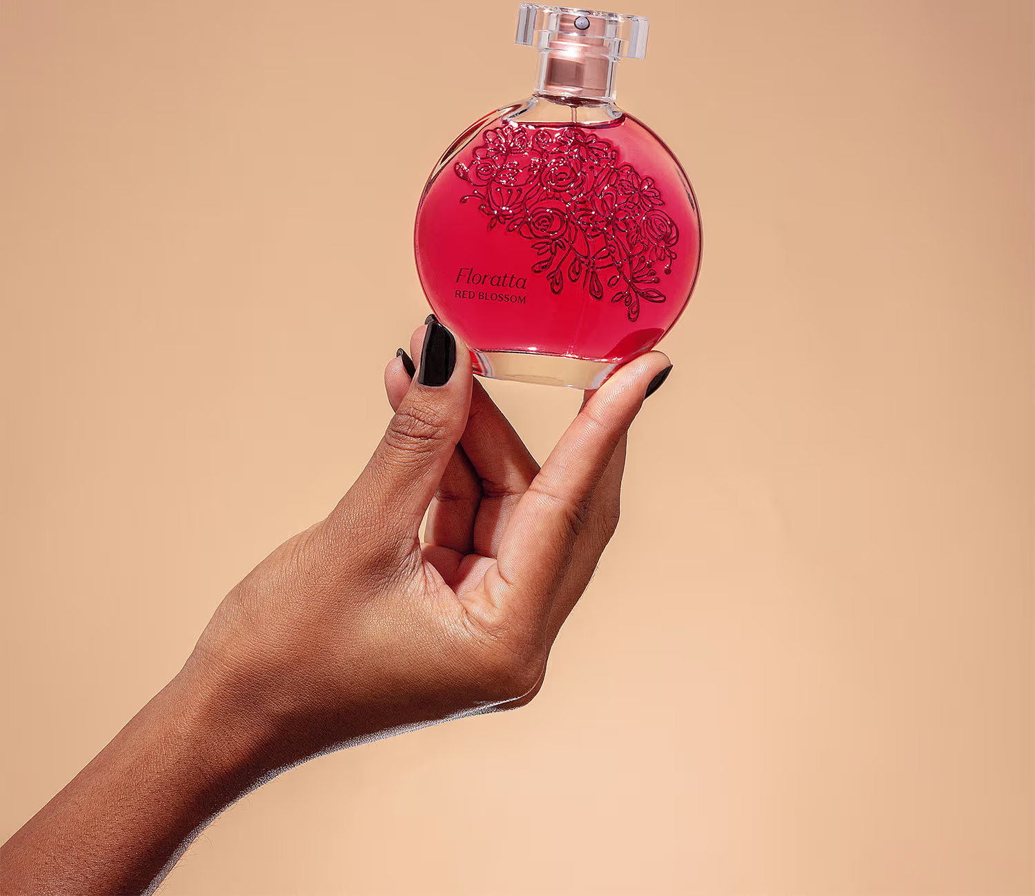 A imagem mostra uma mão segurando um frasco de perfume vermelho com detalhes florais em relevo. O frasco é da marca Floratta e a fragrância se chama Red Blossom (Flor Vermelha, em tradução livre). A mão que o segura tem unhas pintadas de preto.