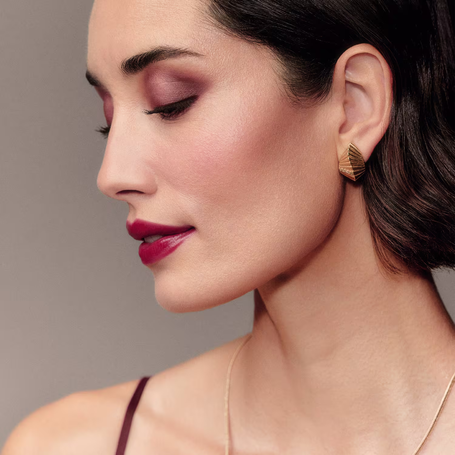 Esta imagem mostra um close-up do rosto de uma mulher usando maquiagem com tons de cereja e cílios longos e curvados. Seus lábios estão pintados com um batom vermelho vivo.