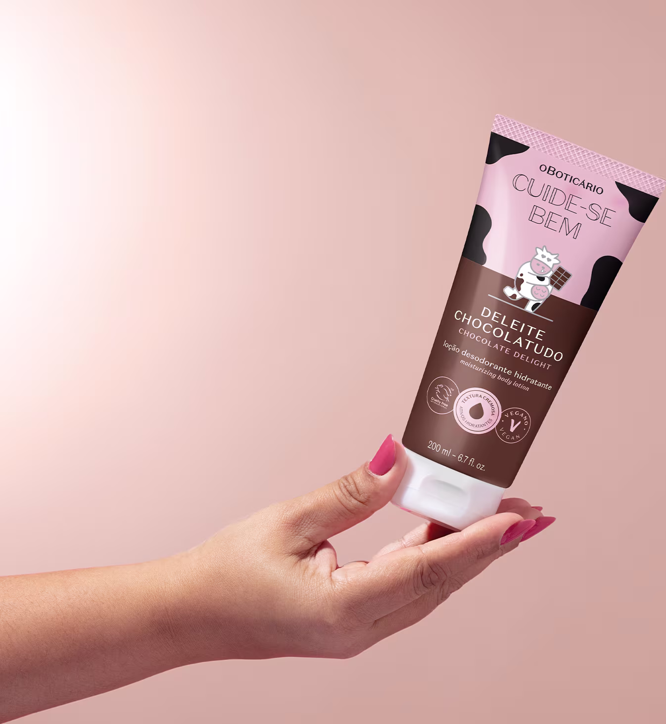 Aqui a loção Cuide-se Bem Deleite Chocolatudo está sendo segurado por uma mão feminina com unhas pintadas de vermelho em fundo rosa claro.