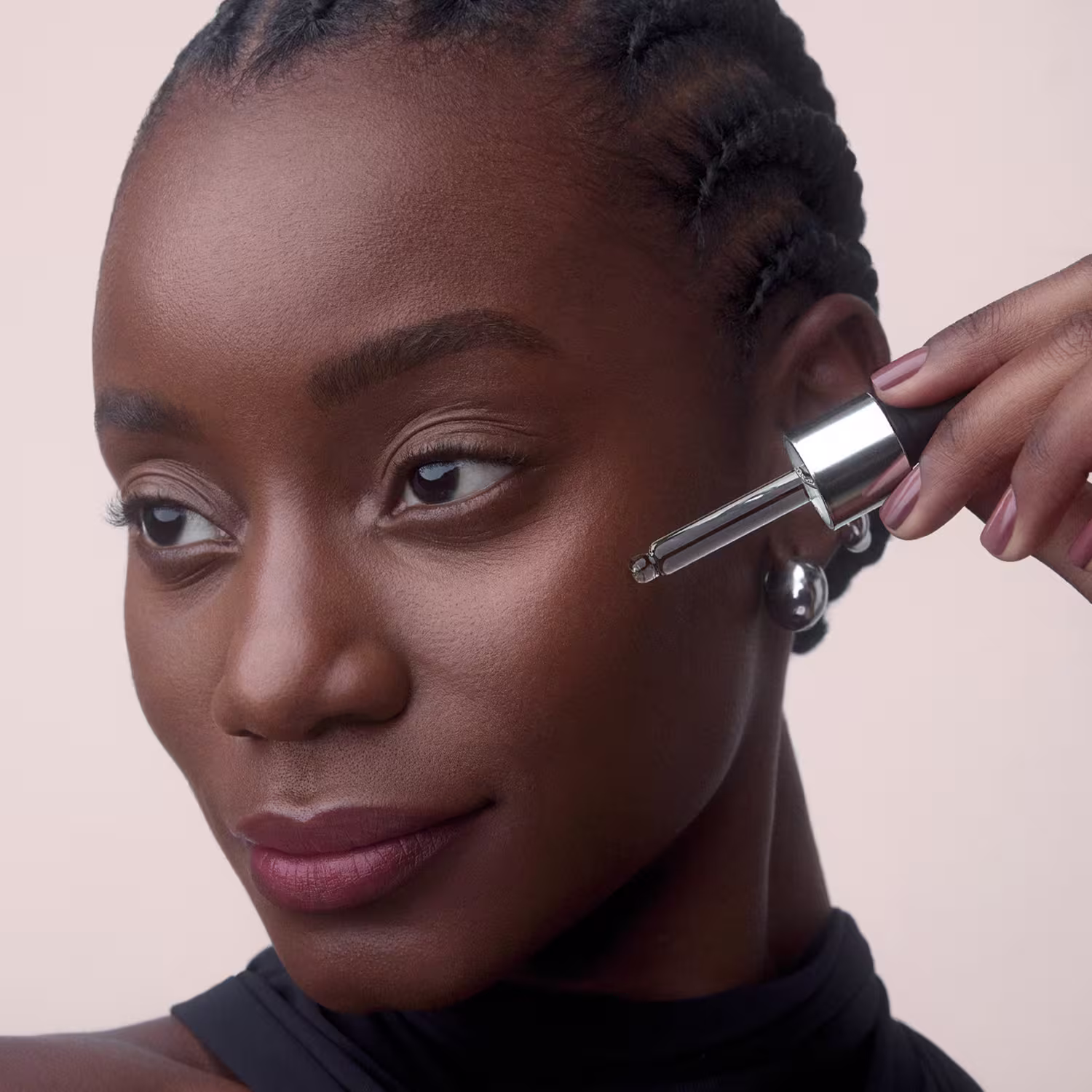 A pessoa na imagem está aplicando um produto de maquiagem em seu rosto. Ela segura um aplicador de maquiagem próximo ao olho, provavelmente aplicando algum tipo de produto para os olhos. A pessoa tem pele escura e cabelo escuro penteado em tranças.