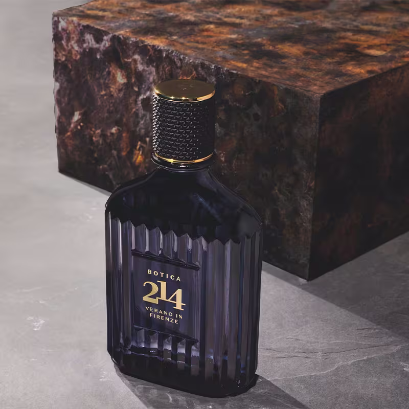 Ambiente elegante e sofisticado, com uma garrafa de fragrância da marca Botica 214, em embalagem escura e dourada, destacada em um fundo de pedra com tonalidades quentes e rústicas, sugerindo um contexto de luxo e exclusividade.
