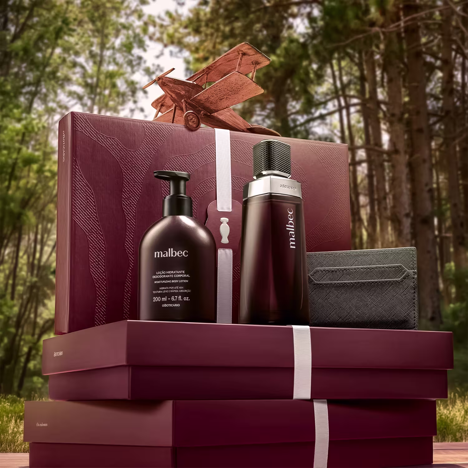 Caixa de presente com produtos de beleza da marca Malbec, em ambiente externo com vegetação ao fundo. Embalagem em tons de vermelho e preto, com desodorante, loção hidratante e perfume, sugerindo um conjunto de cuidados pessoais.