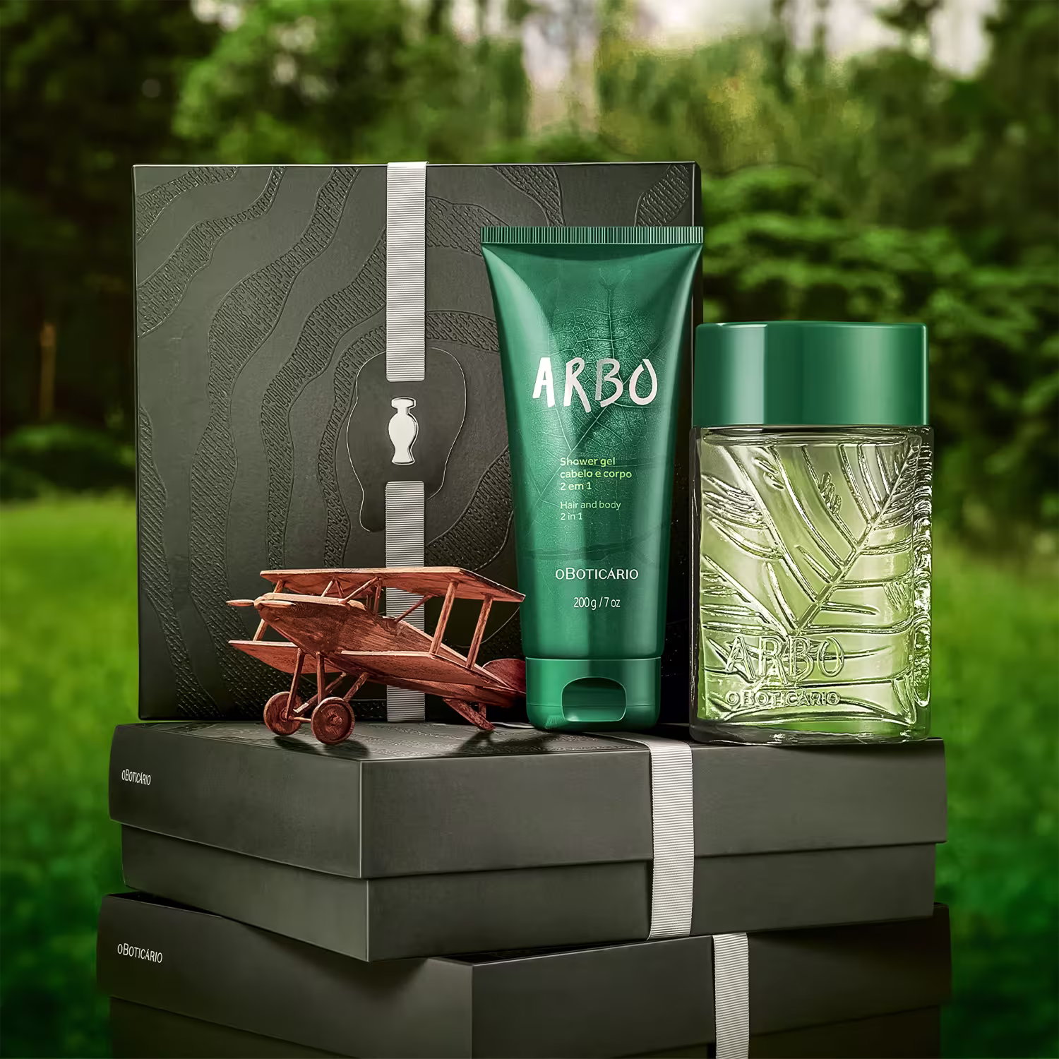 Caixa preta com textura ondulada contendo item de perfumaria da linha Arbo, incluindo frasco de vidro com folhagem e avião de madeira em primeiro plano, sobre superfície cinza em ambiente florestal.