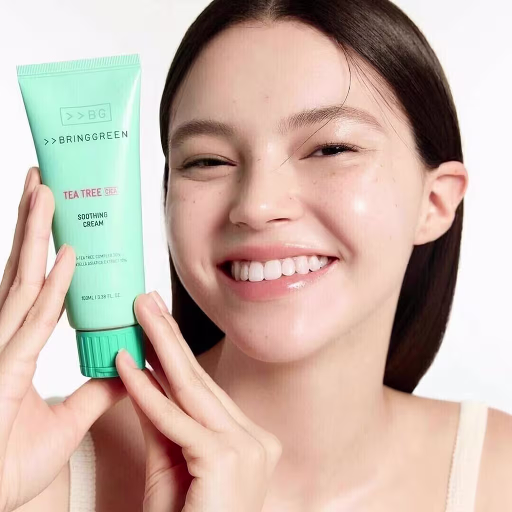 Tubo de creme hidratante para pele da marca Bringgreen, com embalagem verde e rótulo com a descrição "Tea Tree Soothing Cream". O produto é segurado por uma mão feminina sorridente.