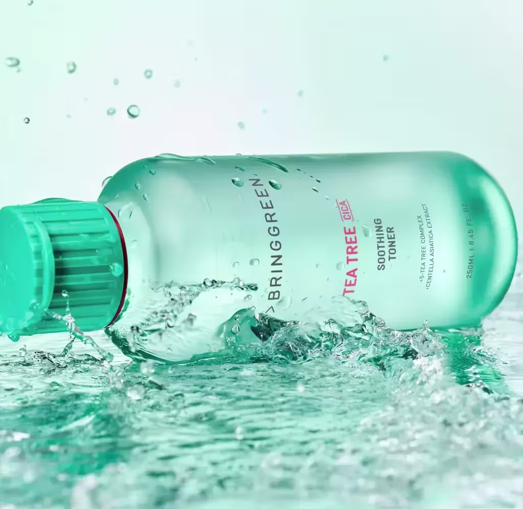 Frasco de plástico verde da marca Bringgreen, contendo um produto de cuidados com a pele chamado "Tea Tree Toning Gel". O frasco está imerso em água, com bolhas e respingos ao redor, criando um efeito visual dinâmico.