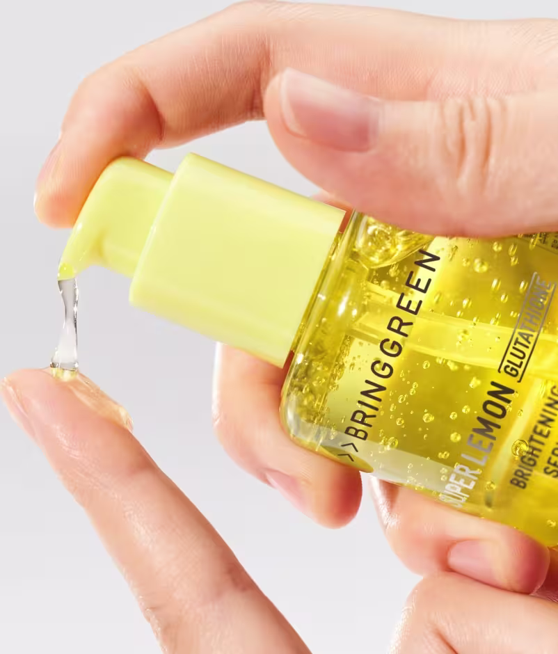 Imagem de um sérum da marca Bringgreen, com líquido amarelo brilhante sendo aplicado em uma mão feminina.